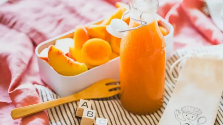 Bílá miska s nakrájenými broskvemi vedle sklenice oranžového sirupu.