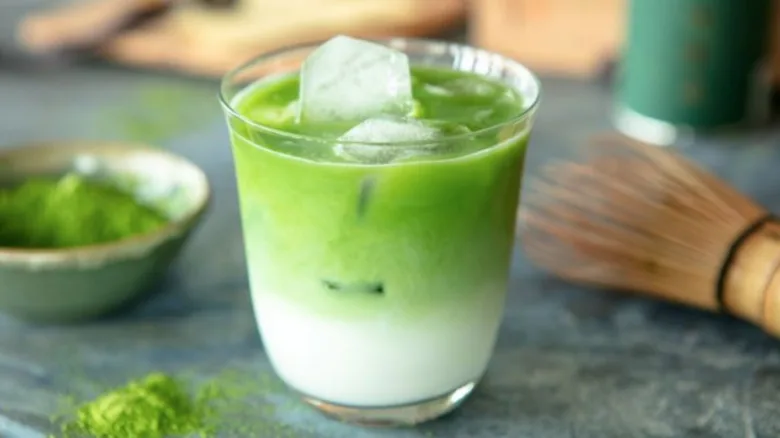 Sklenička s ledem a matcha čajem, vedle bambusový šlehač.