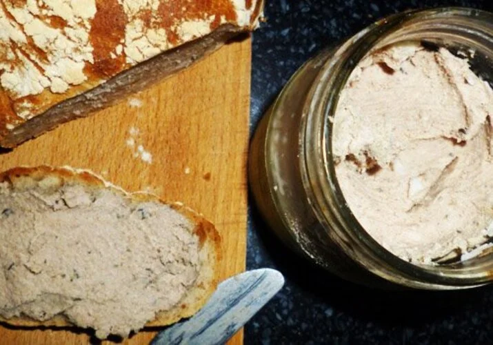 Domácí játrová paštika rozetřená na chlebu vedle sklenice.
