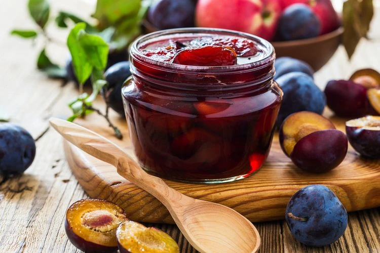 Sklenice domácí švestkové marmelády s čerstvými švestkami na dřevěném talíři.