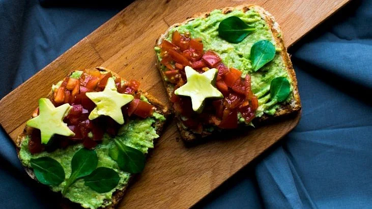 Dva plátky toastu s avokádem, rajčaty a listy špenátu na dřevěném podnose.