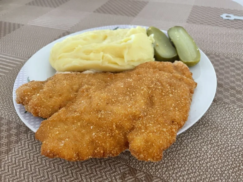 Smažený řízek s bramborovou kaší a okurkovými kousky na talíři.