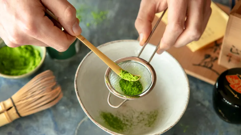 Ruce sítí zelený prášek matcha do čajové misky pomocí bambusového lžičky.