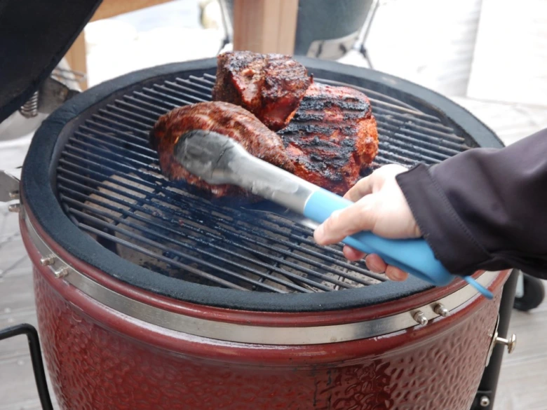 Ruka s kleštěmi upravuje pečené maso na grilu.