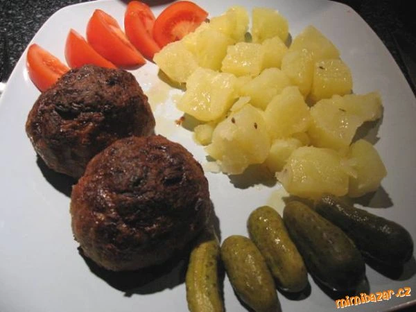 Dva karbanátky s bramborami, rajčaty a okurkami na talíři.