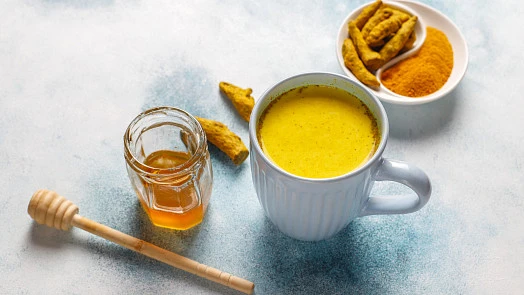 Bílá šálka s kurkumovým latte vedle medu a čerstvého kořene.