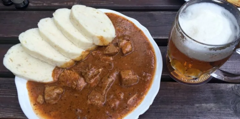 Český guláš s houskovými placičkami a pivem na dřevěném stole.